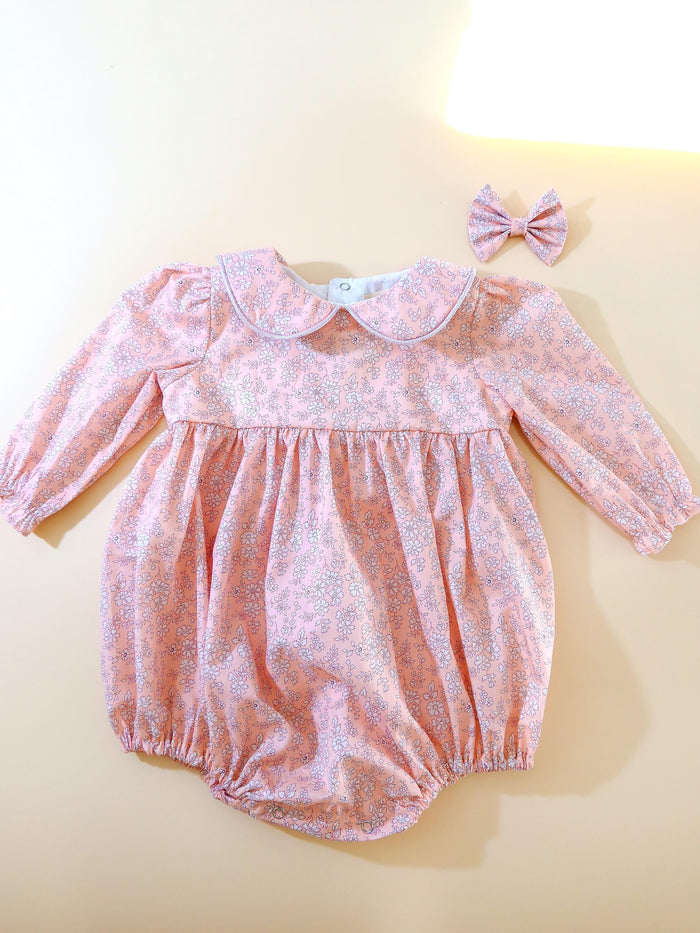 May include: A pink baby romper with a floral pattern and long sleeves, paired with a matching bow. The romper features a Peter Pan collar with a light purple trim. The fabric is a soft pink with a delicate white and gray floral print.