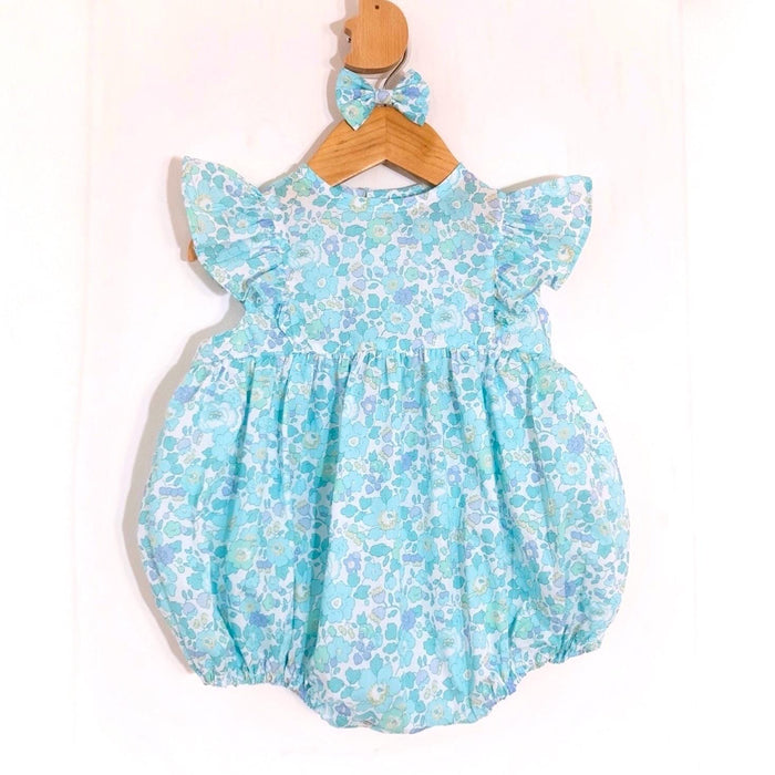 May include: A light blue baby romper with a floral pattern in shades of blue and white. The romper has ruffled sleeves and a matching bow headband. The outfit is hanging on a wooden hanger.
