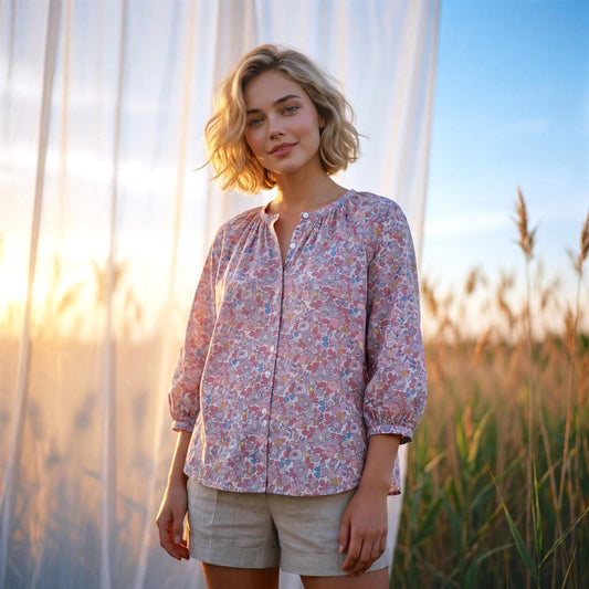 May include: A light pink floral print blouse with three-quarter sleeves and a button-down front. The blouse is paired with beige shorts. The background features tall grass and a sheer white curtain.