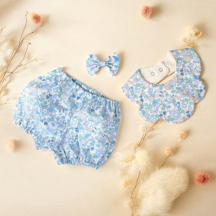 May include: A baby clothing set featuring a pair of bloomers, a bib, and a bow. The items are light blue with a floral pattern in shades of blue and white. The bib has a scalloped edge and two snap closures.