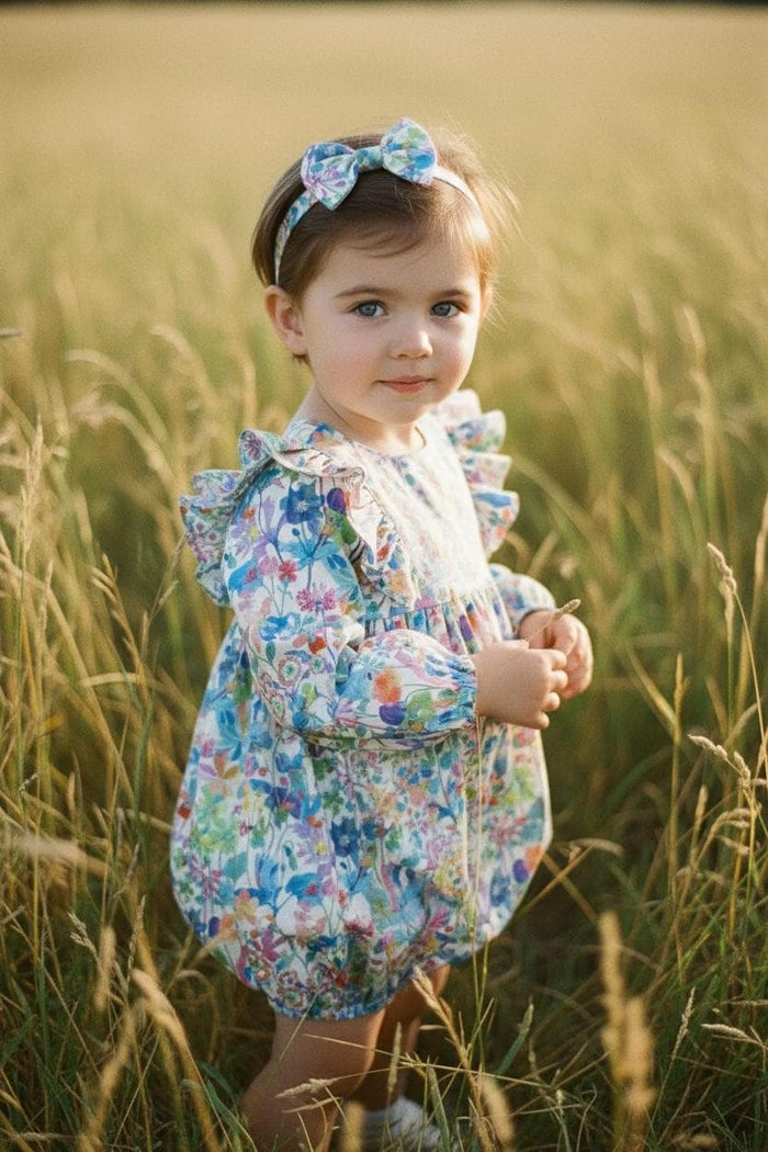 May include: A young child wearing a floral print romper and matching headband stands in a field of tall grass. The romper features long sleeves and a ruffled neckline. The floral pattern includes blue, pink, and green flowers.