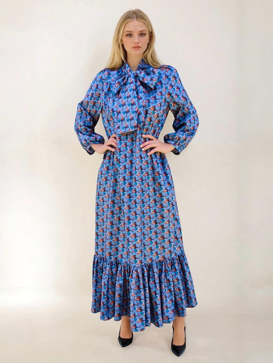 May include: A long, blue dress with a floral pattern. The dress features long sleeves, a large bow at the neckline, and a ruffled hem. The dress is worn with black heels.
