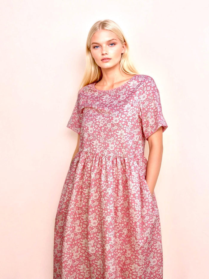 May include: A pink floral print dress with short sleeves and a gathered waist. The dress features a round neckline and pockets. The fabric has a pattern of small white flowers. The model is wearing the dress.