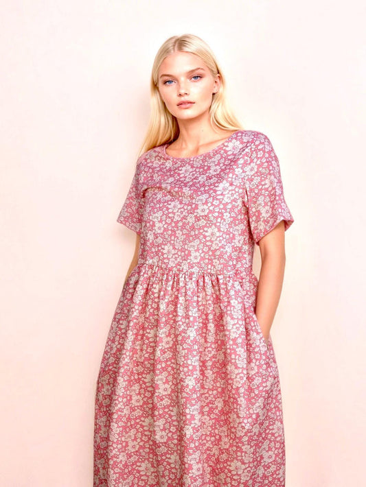 May include: A pink floral print dress with short sleeves and a gathered waist. The dress features a round neckline and pockets. The fabric has a pattern of small white flowers. The model is wearing the dress.