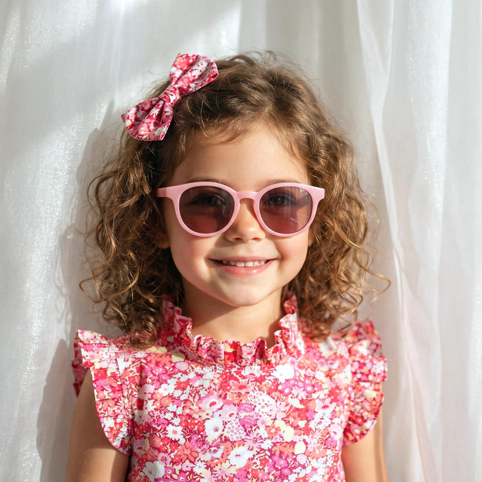 May include: A young girl with curly brown hair wearing pink sunglasses and a matching floral bow. She is also wearing a pink floral dress with a ruffled neckline. The background is a white curtain.