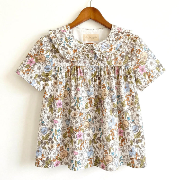 May include: A short-sleeved floral top with a Peter Pan collar. The fabric features a pattern of small flowers in shades of pink, blue, and tan on a white background. The top is hanging on a wooden hanger.