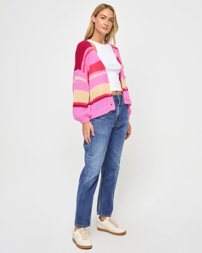 Side view of a model wearing the LSPACE Lido Cardigan in catch some rays, showing the open-knit cardigan with bold horizontal stripes in pink, red, and cream and voluminous sleeves.