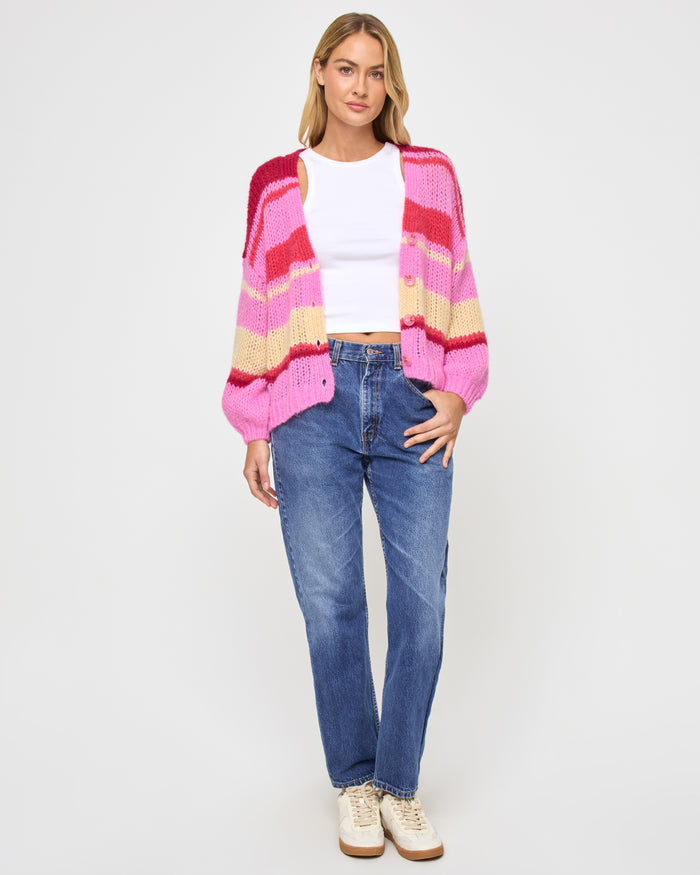 Full-length front view of a model wearing the LSPACE Lido Cardigan in catch some rays, shown open with bold horizontal stripes in pink, red, and cream, styled with jeans and sneakers.