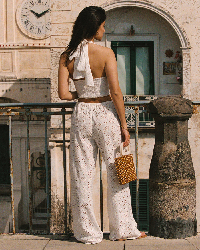 Campaign back view of a model wearing the LSPACE Lily Pant in cream, showing the full-length back of the wide-leg eyelet pants and the matching halter top's tie closure, photographed in a European-style outdoor setting.