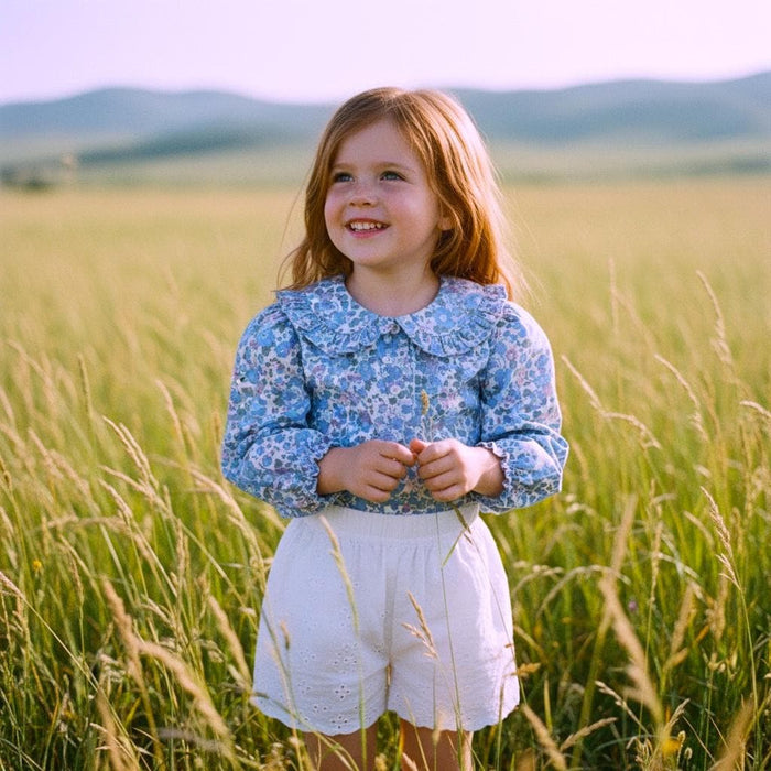 May include: A young girl in a field wearing a blue floral print blouse with a ruffled collar and white eyelet shorts. The blouse has long sleeves and a button-down front. The shorts are high-waisted. The girl has long red hair and is smiling.