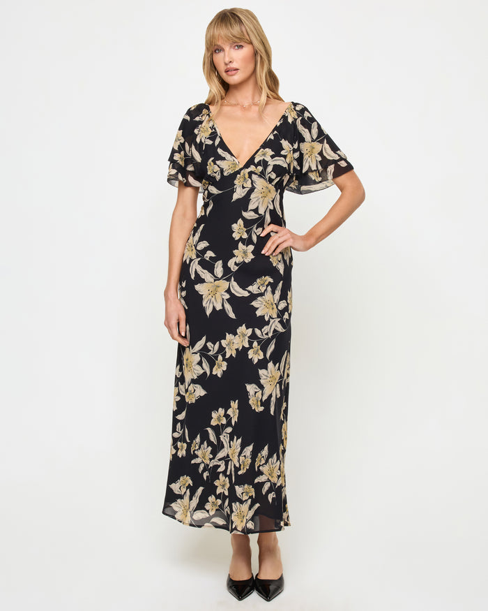 Front view of a model wearing the LSPACE Lover Maxi Dress in petal to the metal, featuring a dark floral print with gold and cream lily blooms, a V-neckline, and flutter short sleeves.