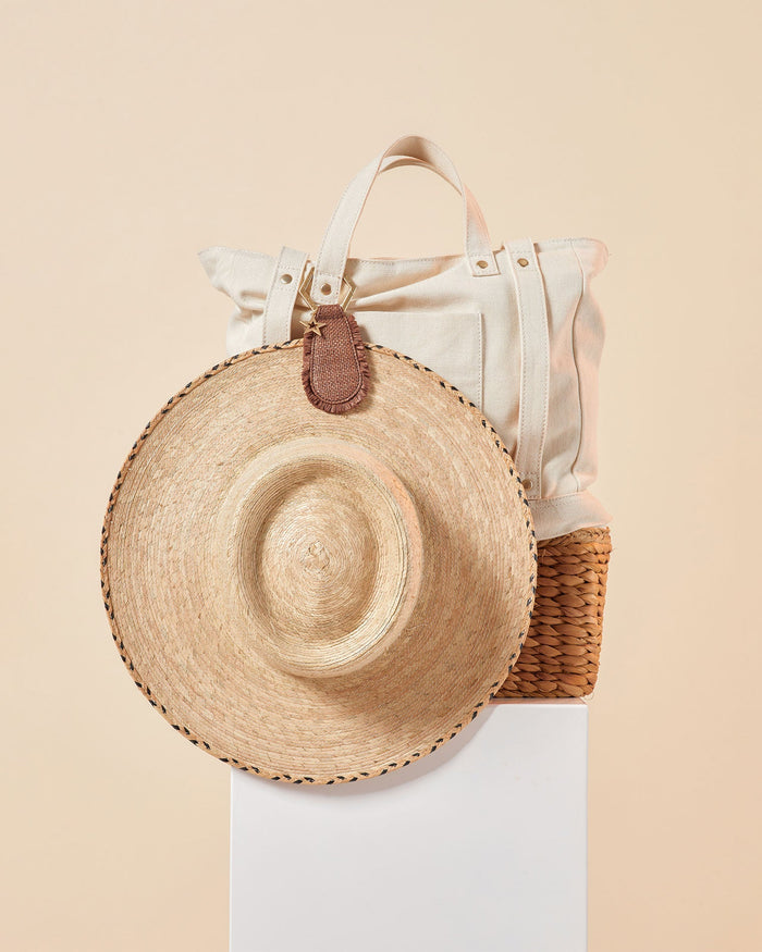 Off-model image of the LSPACE x Lindsay Albanese TOPTOTE clipped to a canvas tote bag alongside a straw hat on a white pedestal.