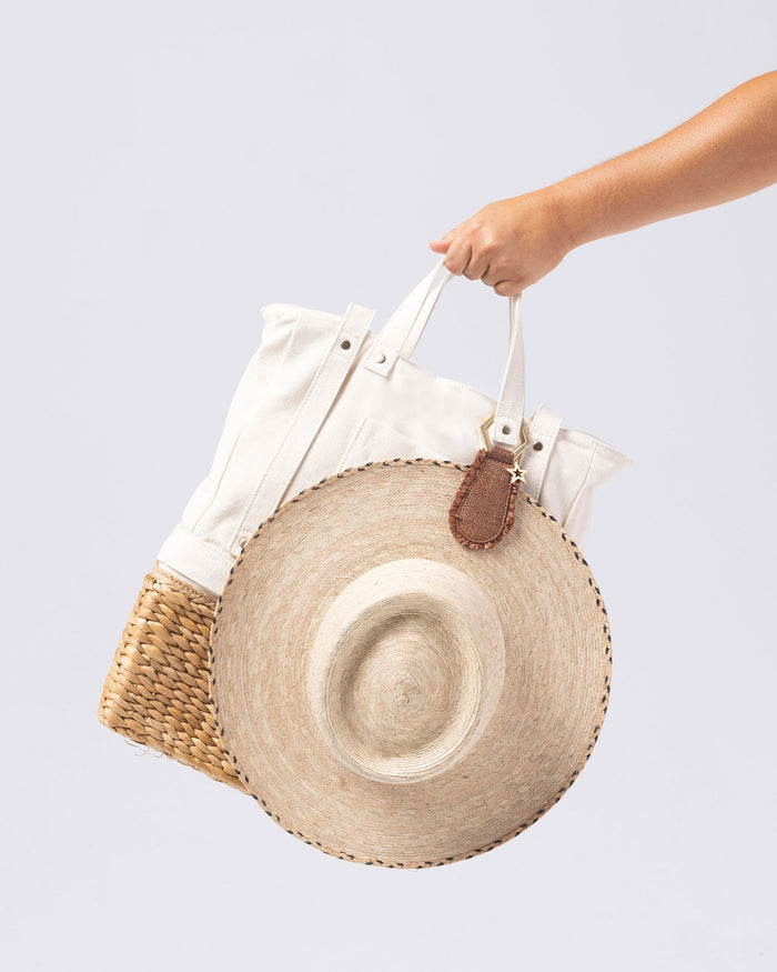 Model's hand holding a canvas tote bag with the LSPACE x Lindsay Albanese TOPTOTE in tobacco attached, alongside a large straw hat.