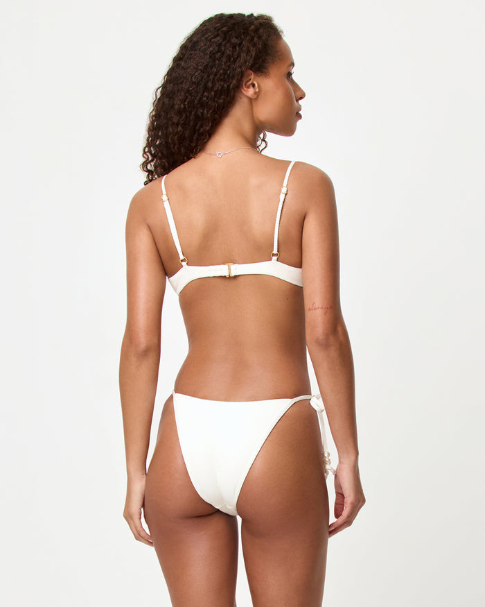 Back view of a model in a studio wearing the LSPACE cream Lucia Bikini Bottom, showing the minimal coverage back and pearl tassel side ties, paired with a cream Sunrise Shell Bikini Top.