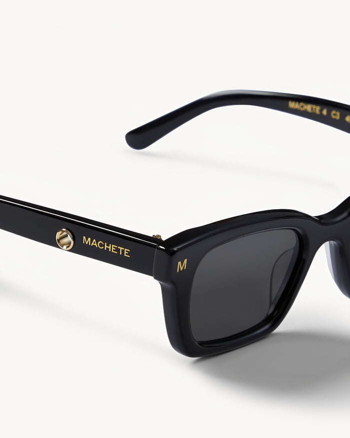 Close-up detail of the Machete Ruby Sunglasses in black showing the Machete branding on the temple arm and dark grey lenses.