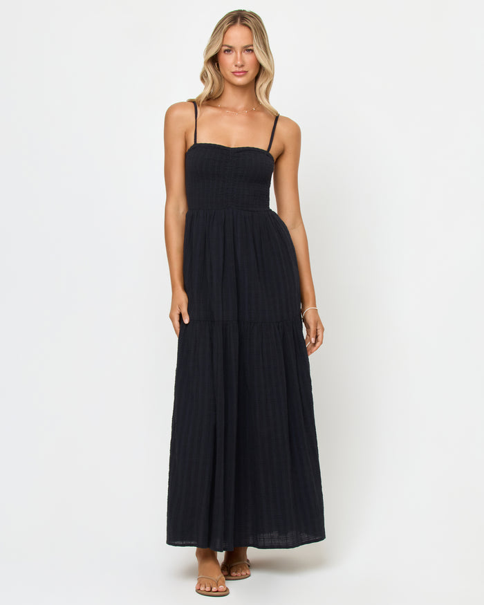 Front view of a model wearing the LSPACE Mallorca Dress in black, a smocked-bodice tiered maxi dress with spaghetti straps, in a textured black fabric, paired with strappy flat sandals.