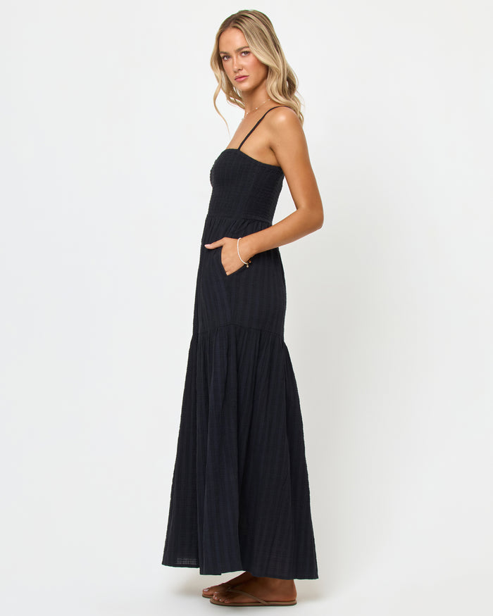 Side view of a model wearing the LSPACE Mallorca Dress in black, showing the smocked bodice and tiered maxi skirt silhouette with a side pocket detail.