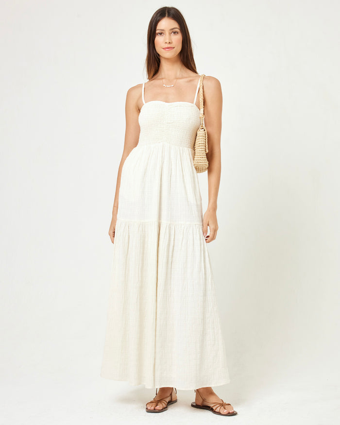 Front view of a model wearing the LSPACE Mallorca Dress in cream, a smocked-bodice tiered maxi dress with spaghetti straps in a cream textured fabric, styled with a woven bag and strappy flat sandals.