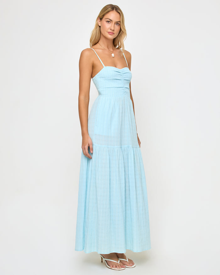Three-quarter front view of a model wearing the LSPACE Mallorca Dress in ibiza blue, a light blue maxi dress with spaghetti straps, a ruched sweetheart bodice, and a tiered flowing skirt.