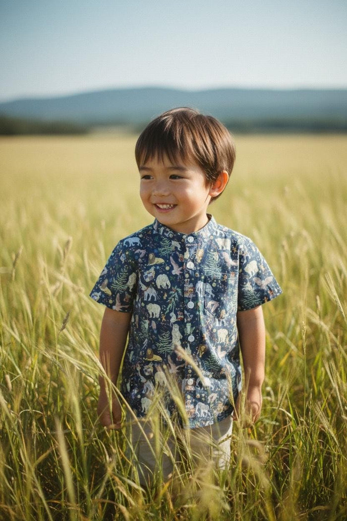 May include: A young child wearing a navy blue short-sleeved shirt with a whimsical forest animal print, standing in a field of tall, golden grass. The shirt has a mandarin collar and button-up front. The child is smiling, with a blurred background of a field and mountains.