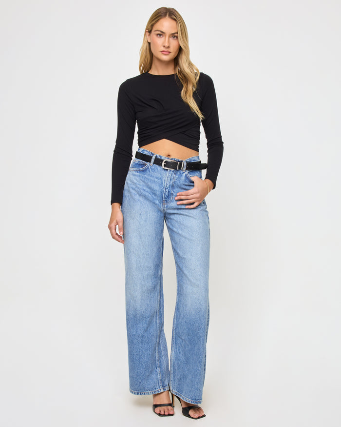 Full-length front view of a model wearing the LSPACE Marseille Top in black, a long-sleeve cropped top with a crew neckline and twisted crossover hem, styled with wide-leg jeans and a black belt.