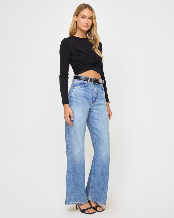 Three-quarter side view of a model wearing the LSPACE Marseille Top in black, a long-sleeve cropped top with a twisted crossover hem, showing the side silhouette.