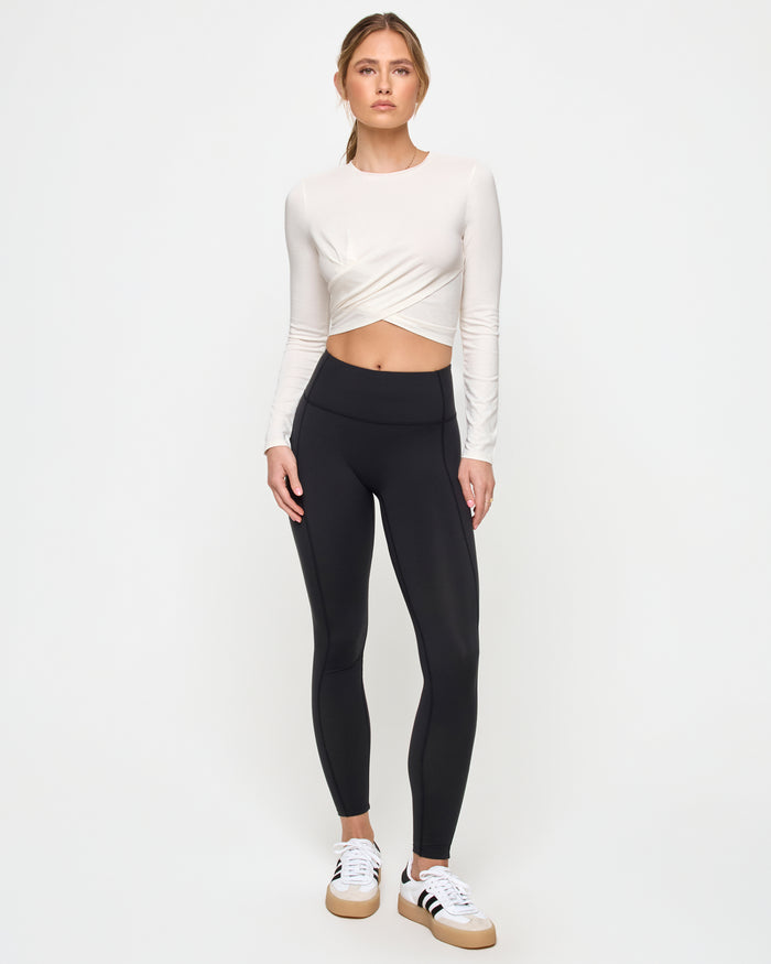 Full-length front view of a model wearing the LSPACE Marseille Top in cream, a long-sleeve cropped top with a crew neckline and twisted crossover hem, styled with black leggings and white sneakers.