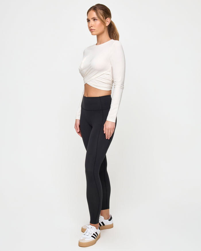 Three-quarter side view of a model wearing the LSPACE Marseille Top in cream, a long-sleeve cropped top with a twisted crossover hem, showing the side silhouette.