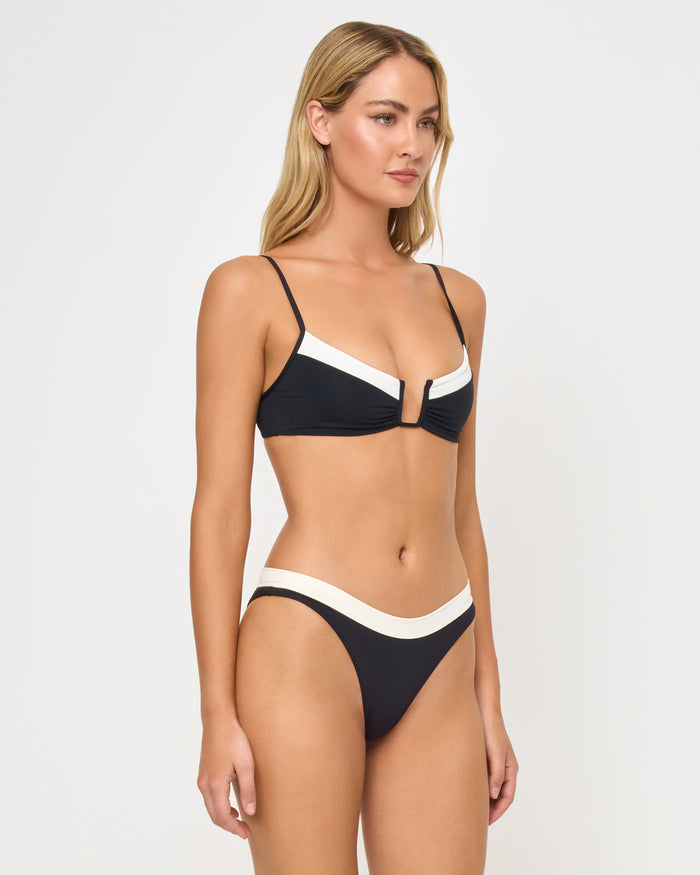 Three-quarter side view of a model wearing the LSPACE black-cream Mercer Bikini Bottom with cream contrast waistband trim, paired with a matching black-cream Peyton Bikini Top.