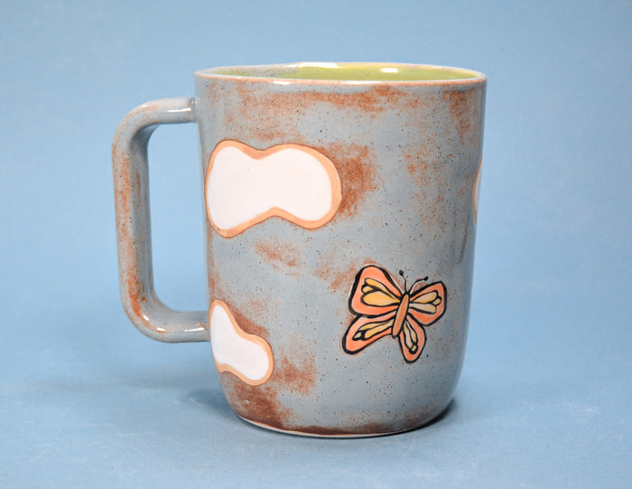 A ceramic mug with a speckled light blue-gray exterior, brown distressed patches, a painted orange butterfly, and two white cloud-like shapes, shown against a solid blue background.