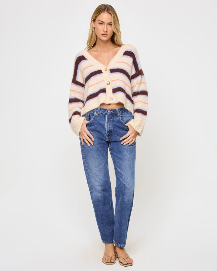 Full-length front view of a model wearing the LSPACE Montauk Cardigan in prep school stripe, a cropped oversized V-neck button-front cardigan in a soft eyelash knit with horizontal stripes in cream, dark brown, lavender, and pink, styled with jeans and sandals.