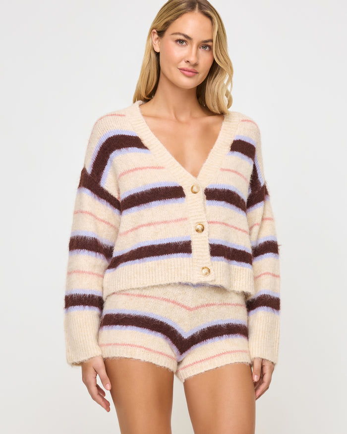 Three-quarter front view of a model wearing the LSPACE Montauk Short in prep school stripe, a high-waisted mini short in a soft eyelash knit with wavy horizontal stripes in cream, dark brown, lavender, and pink.