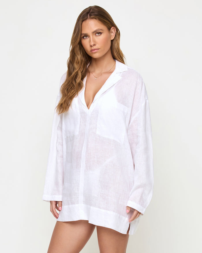 Three-quarter view of a model wearing the LSPACE Oasis Tunic in white, showing the semi-sheer linen fabric, open V-neckline, and long-sleeve oversized relaxed fit.