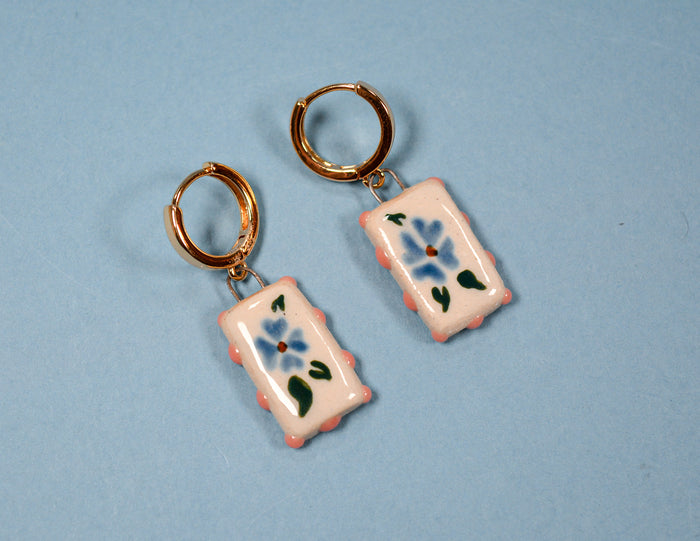 A pair of handmade hoop earrings with rectangular ceramic charms. The charms are light pink with a blue flower, green leaves, and a small red center, hanging from gold-tone hoops against a light blue background.
