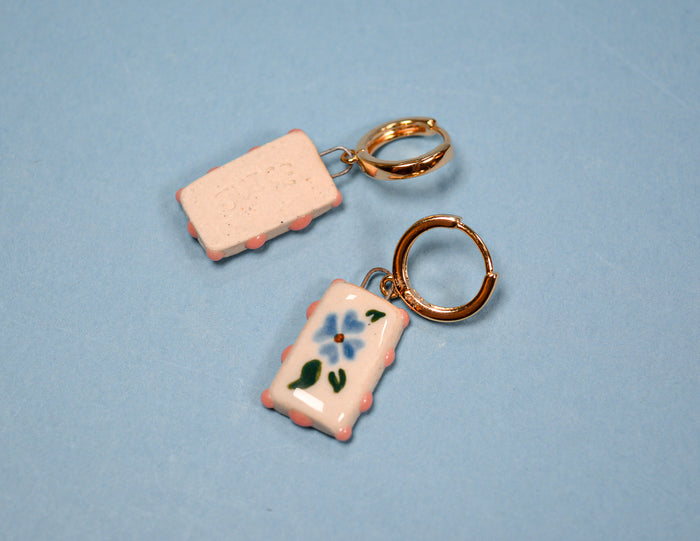 Two small rectangular keychain charms on a light blue background. One charm is pale pink with embossed lettering and a gold key ring.