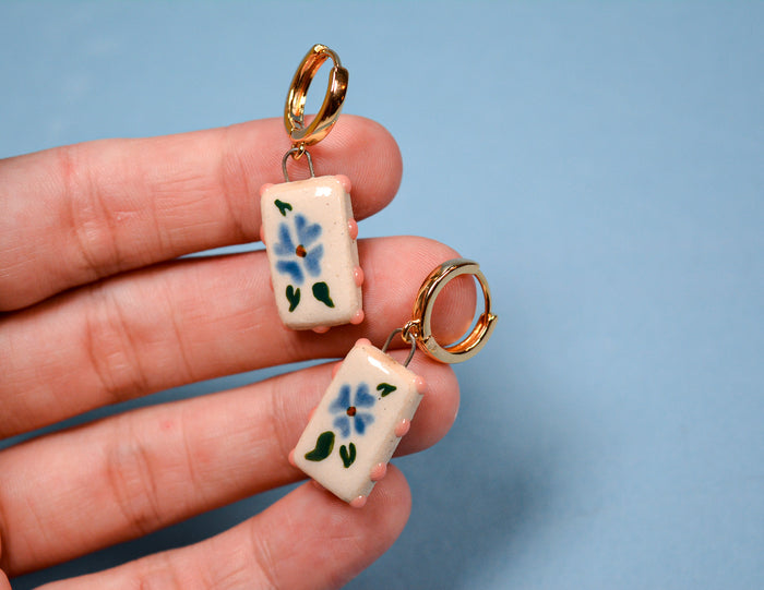A close-up of a hand holding a pair of small gold hoop earrings with rectangular cream-colored charms decorated with blue flowers and green leaves, against a solid light blue background.