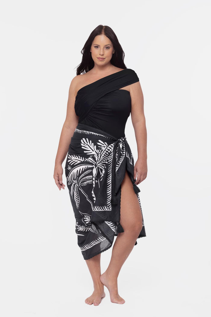 Woman wearing a black one shoulder swimsuit with a palm leaf pattern cover up around the waist