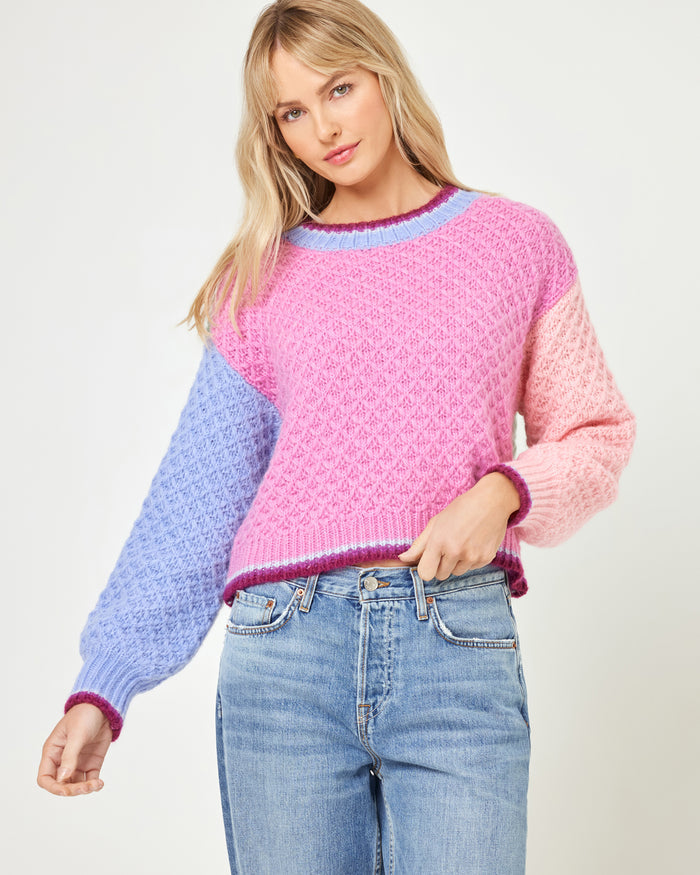 Front three-quarter view of a model wearing the LSPACE Pretty Please Sweater in costa del sol colorblock, featuring a hot pink body, lavender left sleeve, light pink right sleeve, and dark berry trim at the neckline and hem.