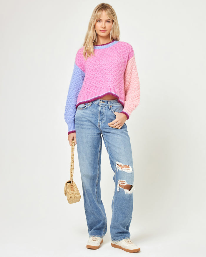 Front full-length view of a model wearing the LSPACE Pretty Please Sweater in costa del sol colorblock, showing the hot pink body, lavender and light pink sleeves, and dark berry trim, styled with distressed blue jeans and white sneakers.