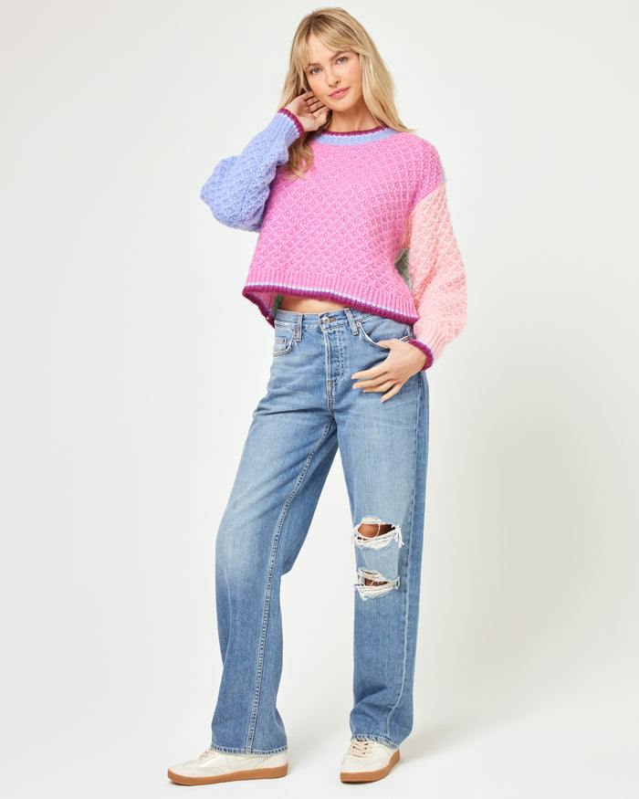 Three-quarter front view of a model wearing the LSPACE Pretty Please Sweater in costa del sol colorblock, featuring the hot pink body and colorblock sleeves, paired with distressed blue jeans and white sneakers.