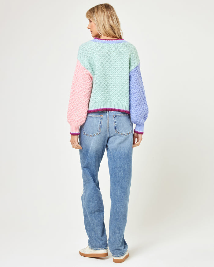 Back view of a model wearing the LSPACE Pretty Please Sweater in costa del sol colorblock, showing the mint green back panel, pink and lavender sleeves, and dark berry trim at the hem and neckline.
