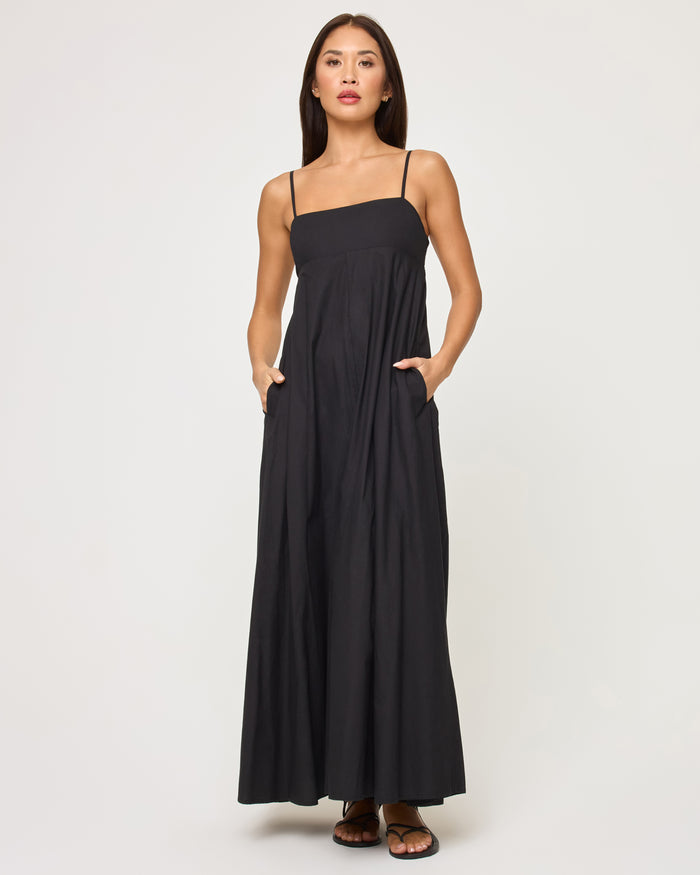 Front view of a model wearing the LSPACE Raveena Dress in black, a relaxed-fit maxi dress with a square neckline, thin spaghetti straps, and a full flowing skirt with side pockets.