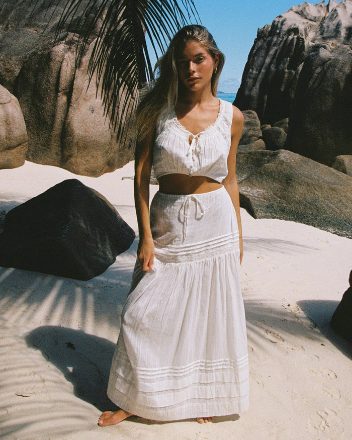Front view of a model wearing the LSPACE Reia Skirt in white, a tiered maxi skirt with pin-tuck and lace trim details, styled with the matching white crop top, photographed on a tropical sandy beach.