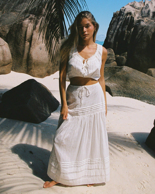 Front view of a model wearing the LSPACE Reia Skirt in white, a tiered maxi skirt with pin-tuck and lace trim details, styled with the matching white crop top, photographed on a tropical sandy beach.
