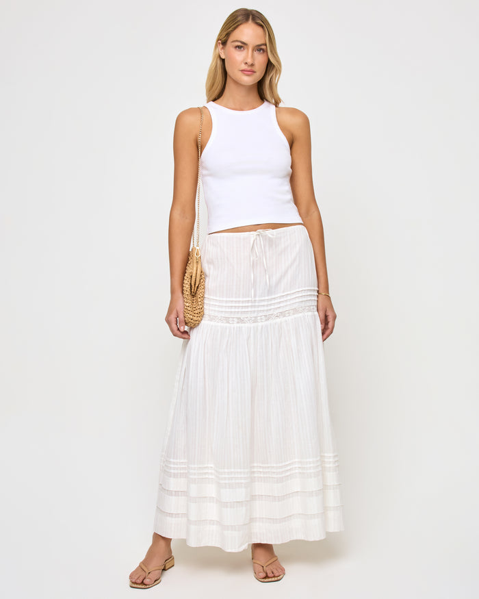 Front view of a model wearing the LSPACE Reia Skirt in white, a tiered maxi skirt with pin-tuck and lace trim details, styled with a white crop top and straw shoulder bag, on a white studio background.