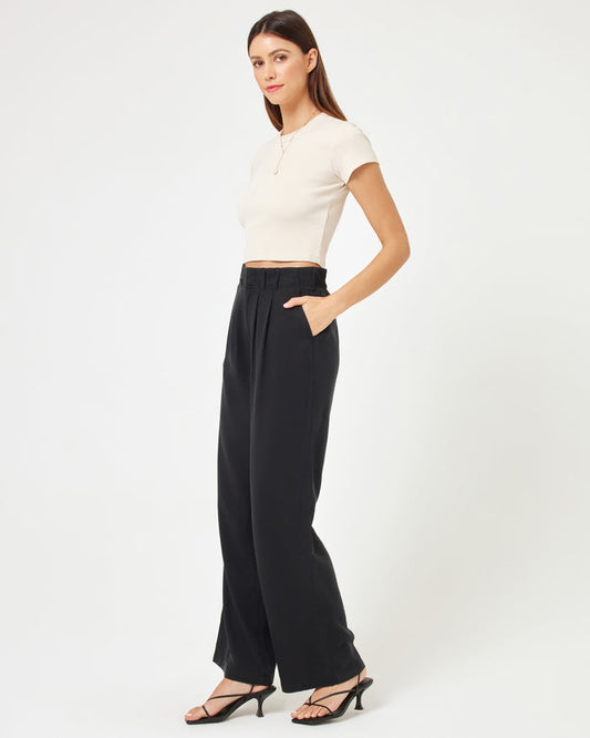 Side view of a model wearing the LSPACE Rhodes Pant in black, a high-waisted wide-leg trouser with front pleats, hand in pocket, styled with a white crop top and strappy heels.