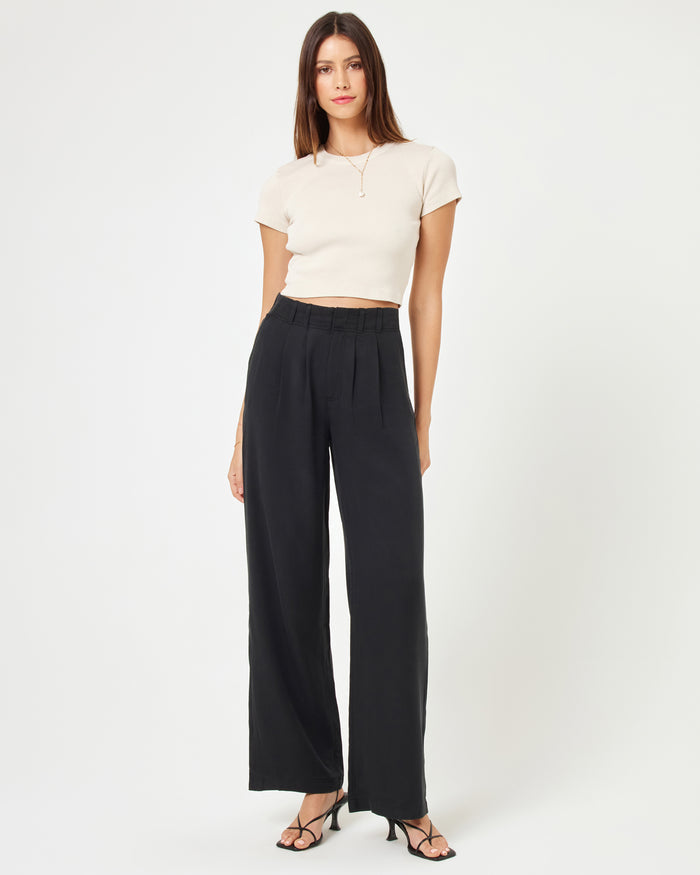 Front view of a model wearing the LSPACE Rhodes Pant in black, featuring a high-waisted wide-leg silhouette with front pleats, styled with a white crop top and strappy heels.
