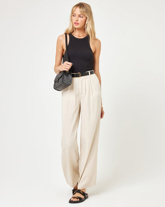 Three-quarter front view of a model wearing the LSPACE Rhodes Pant in oyster, a wide-leg pleated trouser with a black belt, carrying a crossbody bag, styled with a black tank top and sandals.