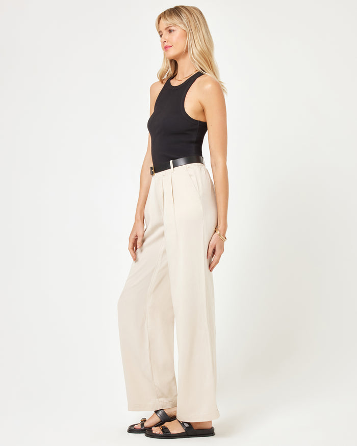 Side view of a model wearing the LSPACE Rhodes Pant in oyster, showing the wide-leg pleated silhouette with black belt detail, styled with a black tank top and sandals.