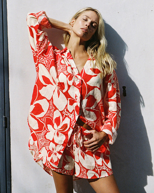 Lifestyle front view of a model wearing the LSPACE Rio Tunic in hibiscus bloom, a long-sleeve button-front shirt in a bold red and white hibiscus print, worn over matching shorts as a beach cover-up.